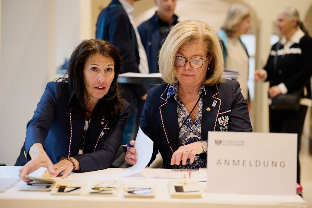 Zwei Frauen sitzen an einem Anmeldeschalter und arbeiten mit Papieren und Dokumenten. Die eine zeigt auf Gegenstände auf dem Tisch, während die andere ein Blatt überprüft. Vor ihnen ist ein Schild mit der Aufschrift ANMELDUNG zu sehen.