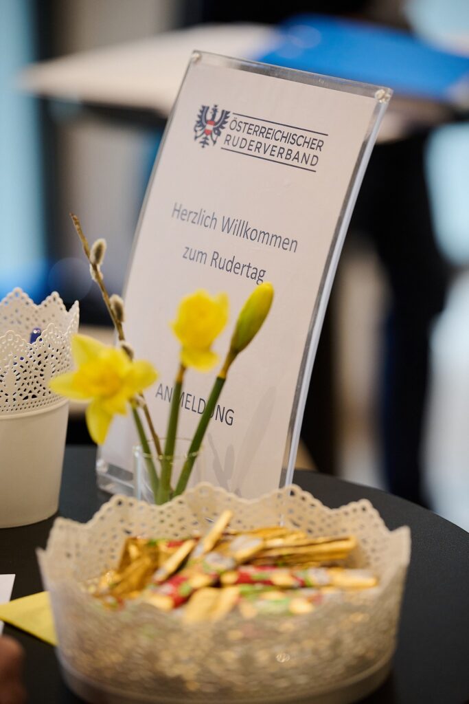 Ein Schild mit der Aufschrift Österreichischer Ruderverband. Herzlich Willkommen zum Rudertag. Die Anmeldung steht auf einem Tisch mit einer Vase mit gelben Blumen und einer Schale mit eingepackten Süßigkeiten im Vordergrund.