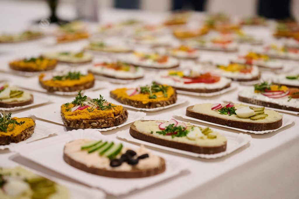 Brotscheiben mit verschiedenen Aufstrichen, Gemüse und Kräutern werden auf weißen Tabletts auf einem Tisch für ein Buffet oder ein Catering angeordnet. Zu den Aufstrichen gehören Radieschen, Gurken, Essiggurken und Grünzeug.