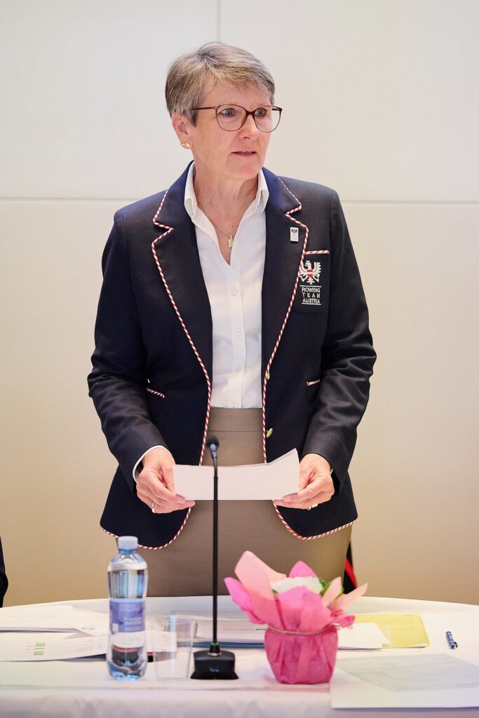 Eine Frau mit Brille und einem marineblauen Blazer mit rotem Rand steht hinter einem Tisch und hält Papiere in der Hand. Auf dem Tisch befinden sich Dokumente, eine Wasserflasche, ein Glas und ein rosa verpackter Gegenstand. Sie scheint zu sprechen oder etwas zu präsentieren.