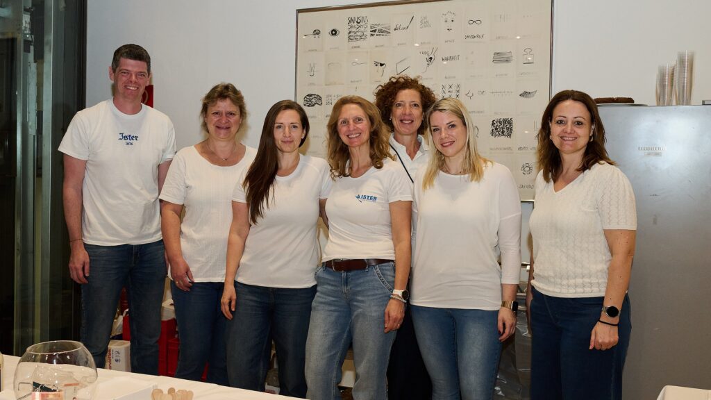 Sieben Erwachsene stehen zusammen in einem Haus und lächeln in die Kamera. Sie alle tragen weiße Oberteile und Jeans. An der Wand hinter ihnen sind Zeichnungen ausgestellt, und im unteren Vordergrund ist ein Tisch mit Glaswaren zu sehen.