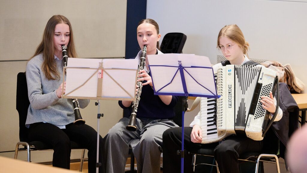 Drei junge Leute sitzen in einem Innenraum und musizieren gemeinsam; zwei spielen Klarinetten, der dritte ein Akkordeon. Vor ihnen stehen Notenständer, und sie scheinen sich auf ihr Spiel zu konzentrieren.