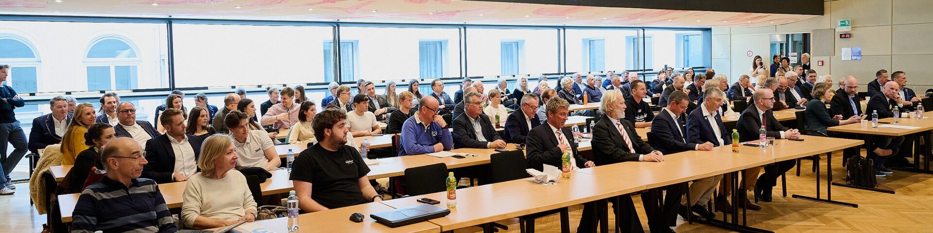 Eine große Gruppe von Menschen, Männer und Frauen, sitzt an langen Tischen in einem modernen Konferenzraum und blickt aufmerksam nach vorne. Einige haben Laptops, Notebooks und Wasserflaschen vor sich stehen.
