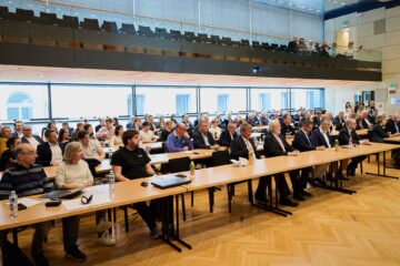 Eine große Gruppe von Menschen, Männer und Frauen, sitzt an langen Tischen in einem modernen Konferenzraum und blickt aufmerksam nach vorne. Einige haben Laptops, Notebooks und Wasserflaschen vor sich stehen.