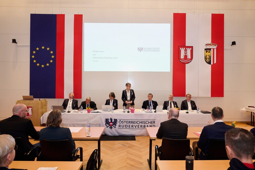 Eine formelle Sitzung mit acht Personen, die an einem langen Tisch sitzen, und einer Person, die steht, vor großen Flaggen der EU, Österreichs und einer Region, wobei eine Präsentation an die Wand projiziert wird.