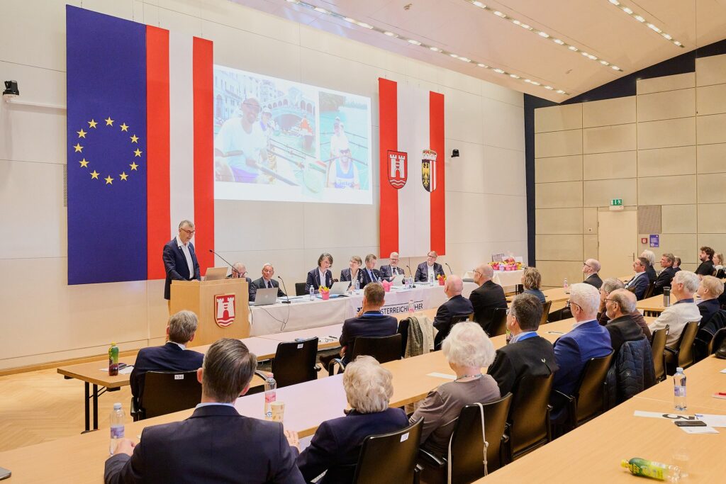 Eine formelle Veranstaltung in einem Konferenzraum, bei der die Teilnehmer gegenüber einer Bühne sitzen, auf der die Redner an einem langen Tisch Platz genommen haben. Große Flaggen der Europäischen Union und der Regionen hängen an der Wand, und eine Präsentation wird auf eine Leinwand projiziert.