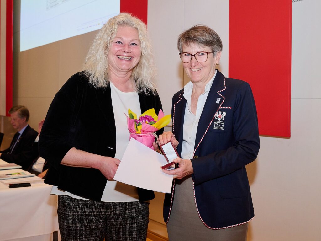 Zwei Frauen lächeln bei einer feierlichen Veranstaltung; die eine hält Blumen und eine Urkunde, die andere trägt einen marineblauen Blazer mit Abzeichen und Medaille und steht vor einer weißen Wand mit roten Paneelen. Im Hintergrund sitzen Menschen an Tischen.