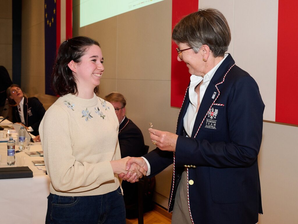 Eine junge Frau in einem cremefarbenen Pullover schüttelt einer älteren Frau in einem marineblauen Blazer bei einer Indoor-Veranstaltung die Hand und lächelt, während andere an einem Tisch im Hintergrund sitzen.