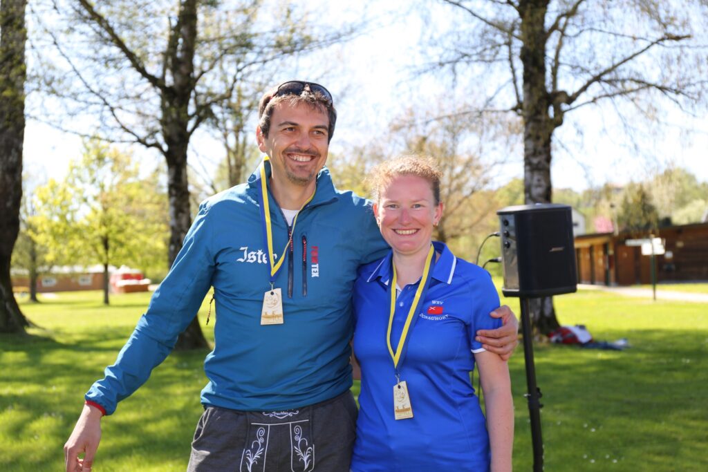 Zwei Personen stehen lächelnd im Freien und tragen Medaillen an gelben Bändern um den Hals. Der Mann auf der linken Seite hat seinen Arm um die Frau auf der rechten Seite gelegt. Im Hintergrund sind Bäume und Gras zu sehen.