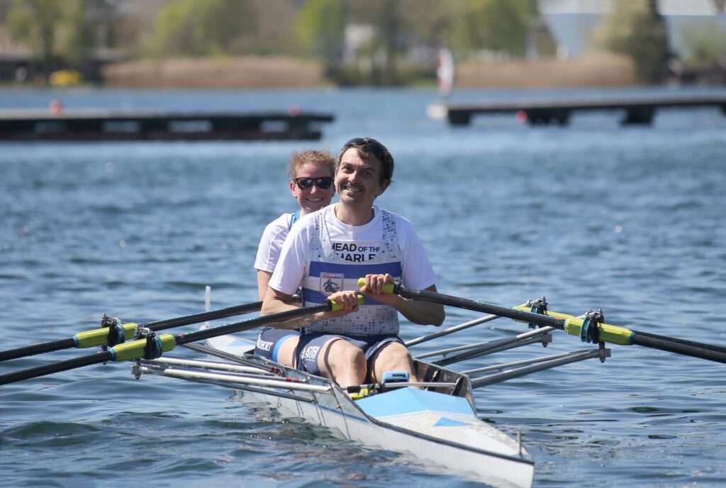 Zwei Männer in weißen Hemden rudern gemeinsam in einem Doppelzweierboot auf ruhigem Wasser, lächeln und genießen die Aktivität an einem sonnigen Tag. Im Hintergrund sind Docks und Bäume zu sehen.