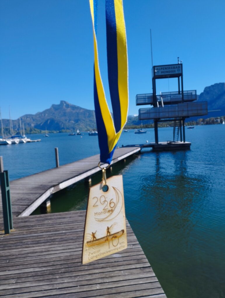 Eine Goldmedaille mit blau-gelber Schleife hängt vor einem See. Im Hintergrund sind Segelboote, ein hölzerner Steg, ein Sprungturm mit der Aufschrift Alpenseebad Mondsee und ferne Berge unter einem klaren blauen Himmel zu sehen.