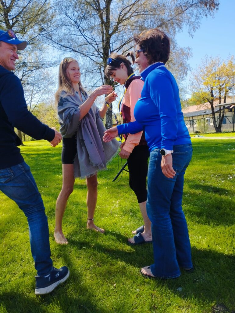 Vier Erwachsene stehen zusammen auf einer sonnigen Wiese mit Bäumen im Hintergrund. Eine Frau ist barfuß, lächelt und hält einer anderen Frau etwas hin. Alle wirken fröhlich und in ein Gespräch vertieft.