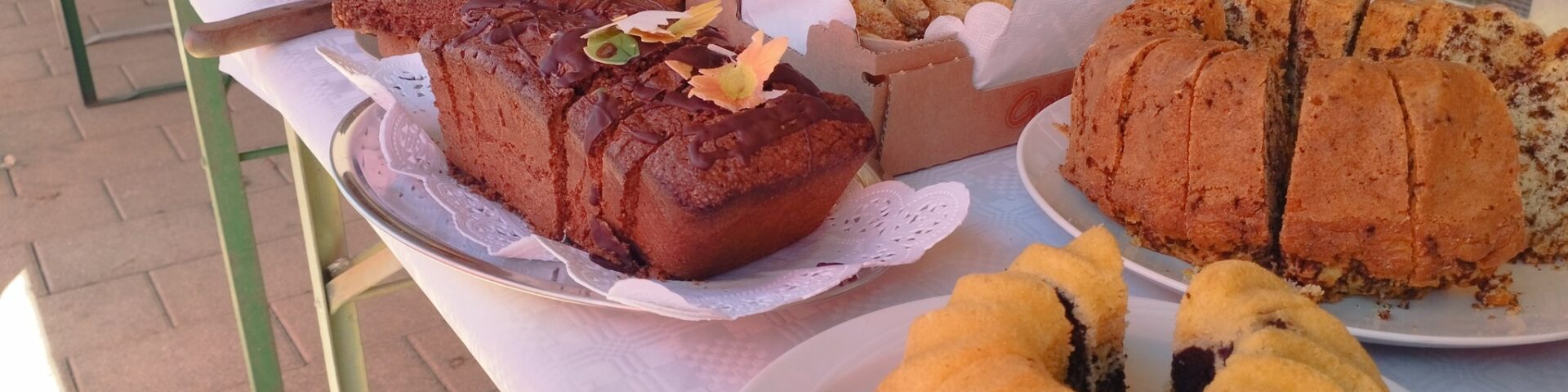 Ein langer Tisch mit verschiedenen Kuchen und Gebäckstücken, darunter Gugelhupf, Laibkuchen und Kekstabletts, die an einem sonnigen Tag im Freien neben einem Metallzaun ausgestellt sind.