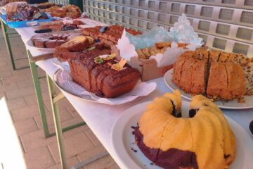 Ein langer Tisch mit verschiedenen Kuchen und Gebäckstücken, darunter Gugelhupf, Laibkuchen und Kekstabletts, die an einem sonnigen Tag im Freien neben einem Metallzaun ausgestellt sind.