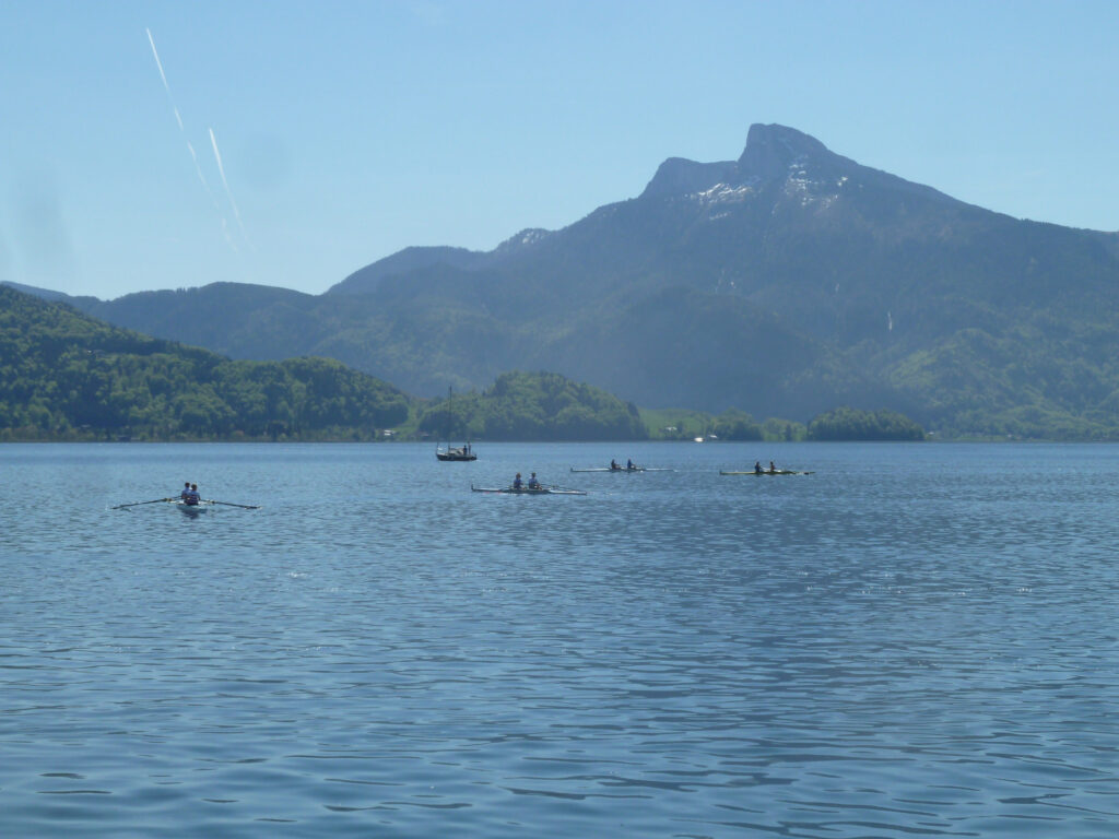 Menschen rudern in kleinen Booten auf einem ruhigen See mit sanftem Wellengang, umgeben von grünen Hügeln und einem hohen Berg im Hintergrund unter einem klaren blauen Himmel.