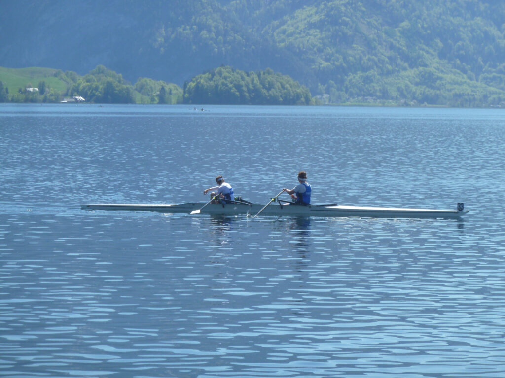 Zwei Personen rudern in einem schmalen Boot auf einem ruhigen See, der von grünen Hügeln und Bergen umgeben ist, unter einem klaren Himmel.