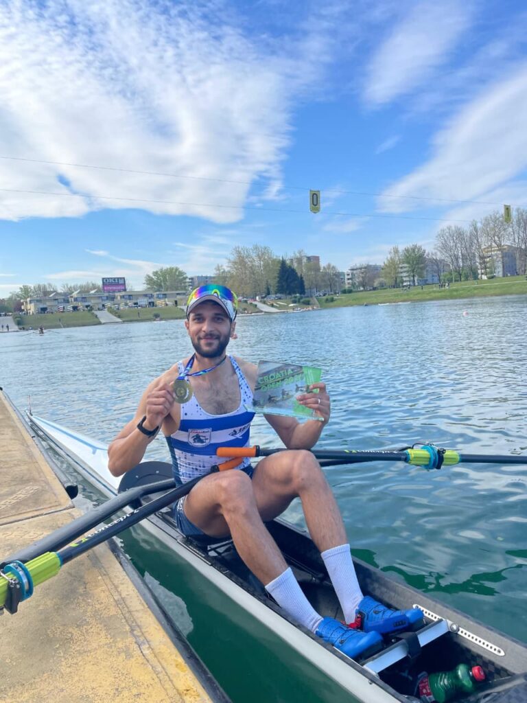 Ein männlicher Ruderer sitzt in einem Ruderboot auf einem See, trägt Sportkleidung und eine Sonnenbrille, lächelt und hält eine Medaille und eine Urkunde in der Hand, im Hintergrund blauer Himmel, Wasser und Grün.